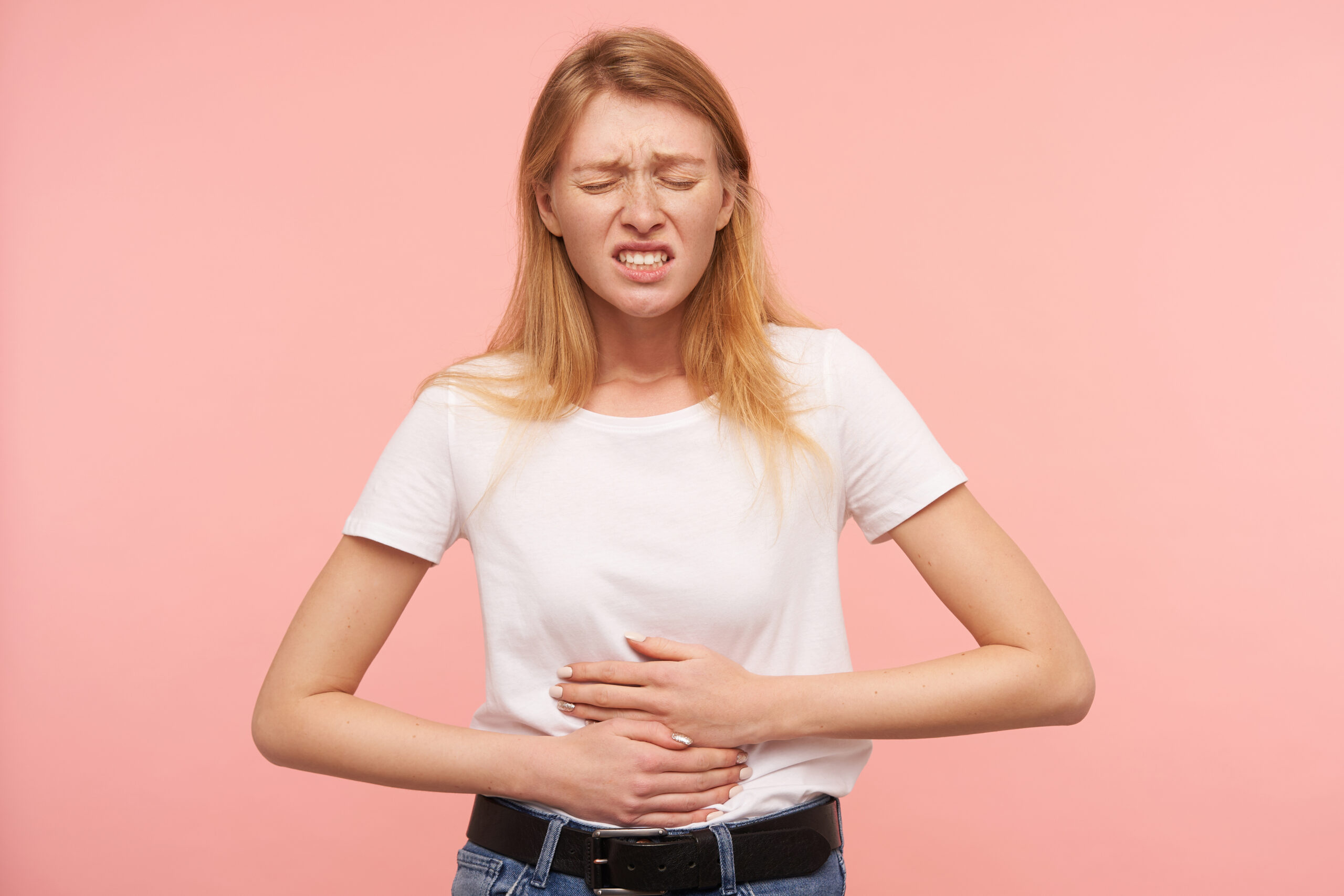 Indoor photo of young pretty redhead lady keeping her eyes closed and frowning face while having stomachache, standing over pink background in casual clothes Indoor photo of young pretty redhead lady keeping her eyes closed and frowning face while having stomachache, standing over pink background in casual clothes