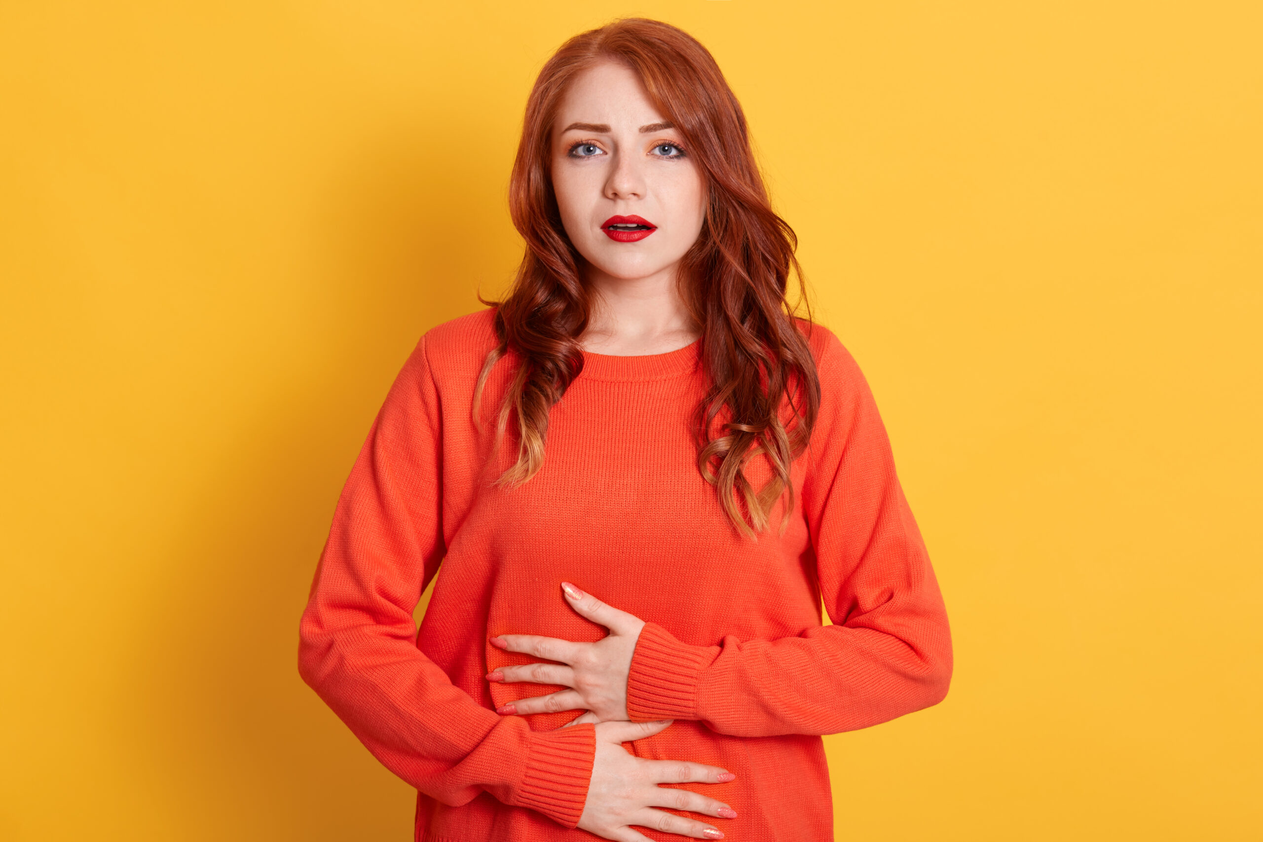 Woman with stomachache, having food poisoning, posing isolated over yellow background, looks at camera, lady has red wavy hair wearing casual orange sweater. Woman with stomachache, having food poisoning, posing isolated over yellow background, looks at camera, lady has red wavy hair wearing casual orange sweater.