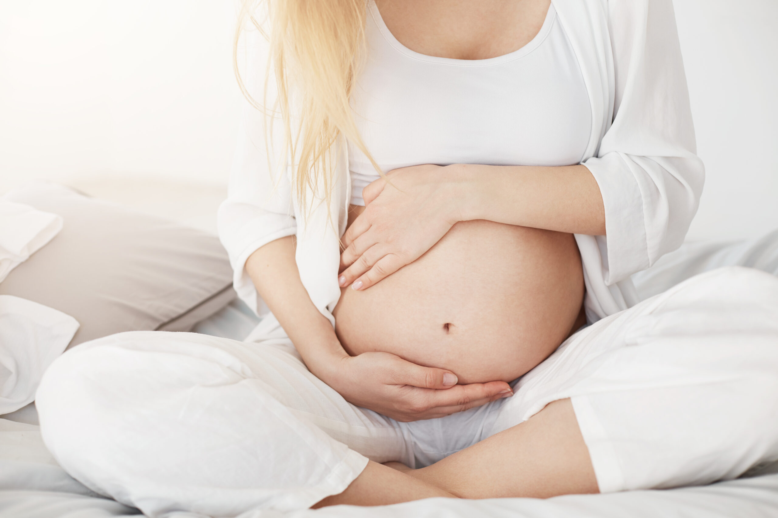 Young pregnant soon to be mother touching her belly sitting in her bedroom in afternoon. Pregnancy concept. High key. Young pregnant soon to be mother touching her belly sitting in her bedroom in afternoon. Pregnancy concept. High key.