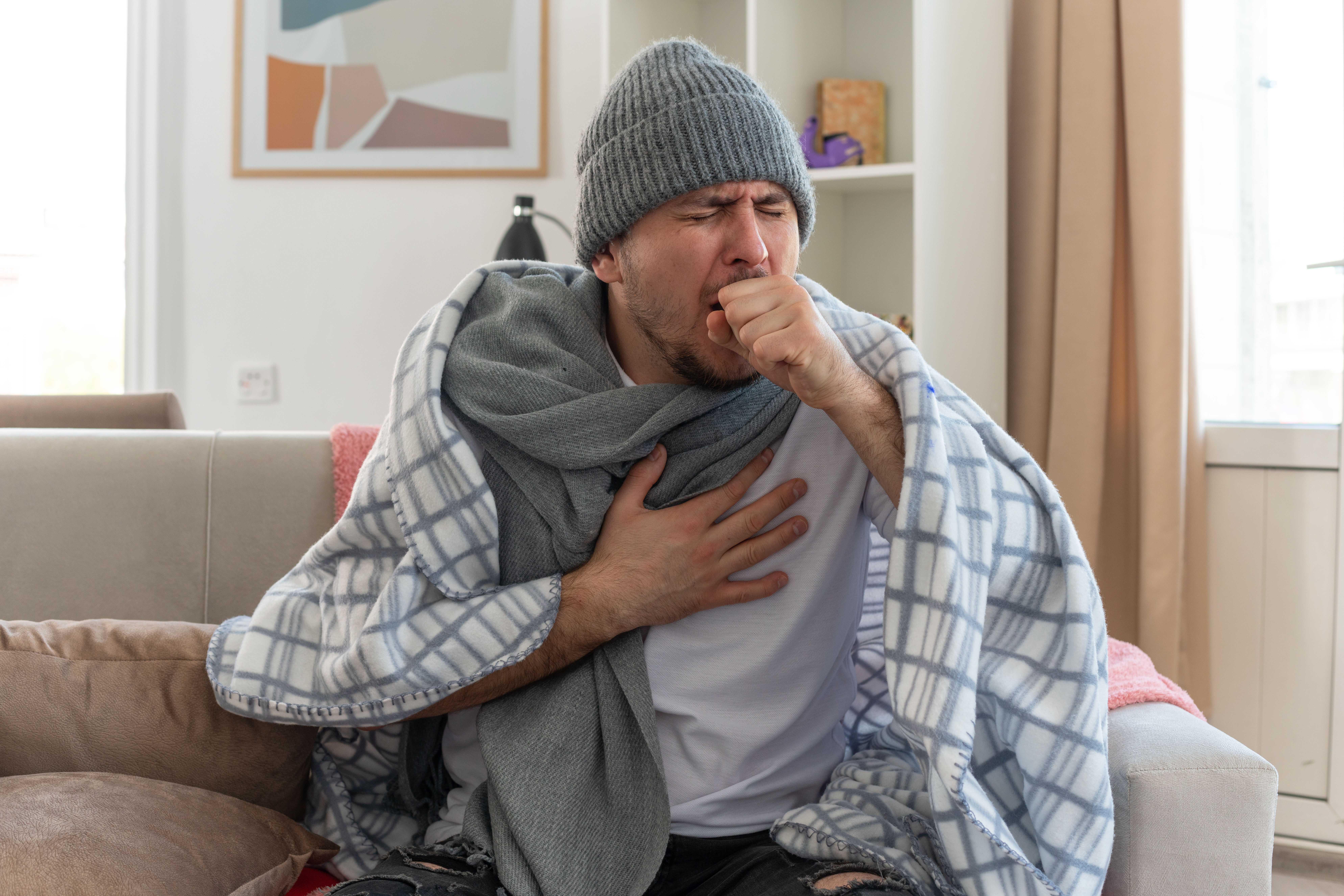 aching ill slavic man with scarf around neck wearing winter hat wrapped in plaid coughing keeping fist close to mouth sitting on couch at living room aching ill slavic man with scarf around neck wearing winter hat wrapped in plaid coughing keeping fist close to mouth sitting on couch at living room
