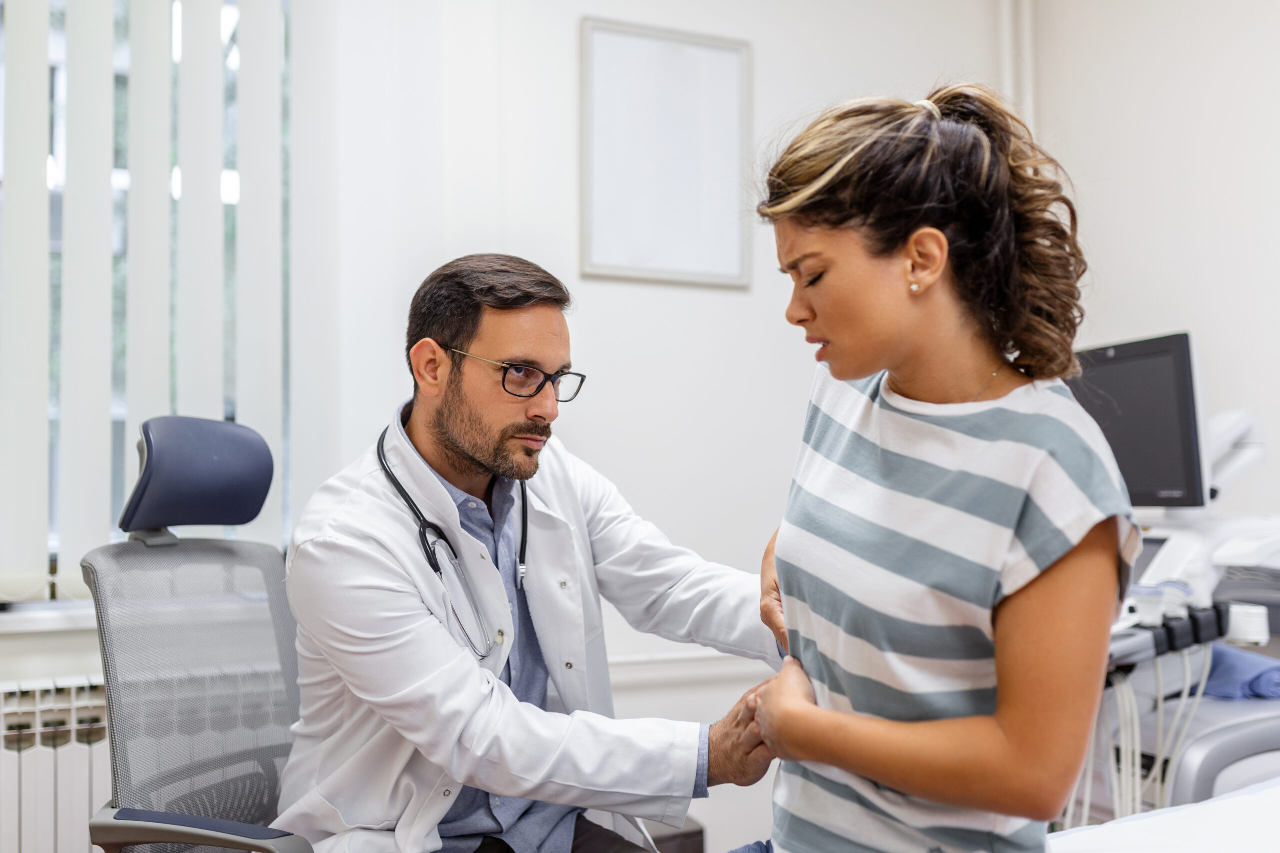 Patient telling physician about her pain and health problems during visit to hospital. Young woman complaining about back or kidney ache while sitting on examination bed at the doctor’s office Patient telling physician about her pain and health problems during visit to hospital. Young woman complaining about back or kidney ache while sitting on examination bed at the doctor’s office