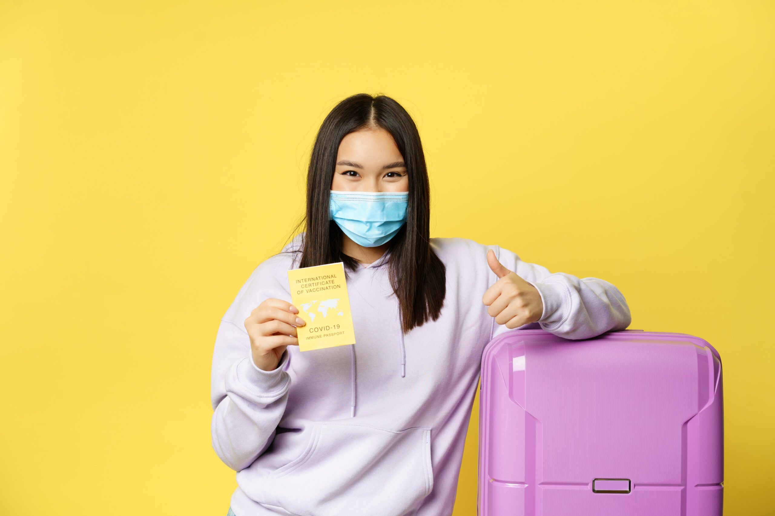 Smiling asian girl tourist in face mask, standing with suitcase, showing covid international vaccination certificate for travellers and thumbs up, yellow background Smiling asian girl tourist in face mask, standing with suitcase, showing covid international vaccination certificate for travellers and thumbs up, yellow background