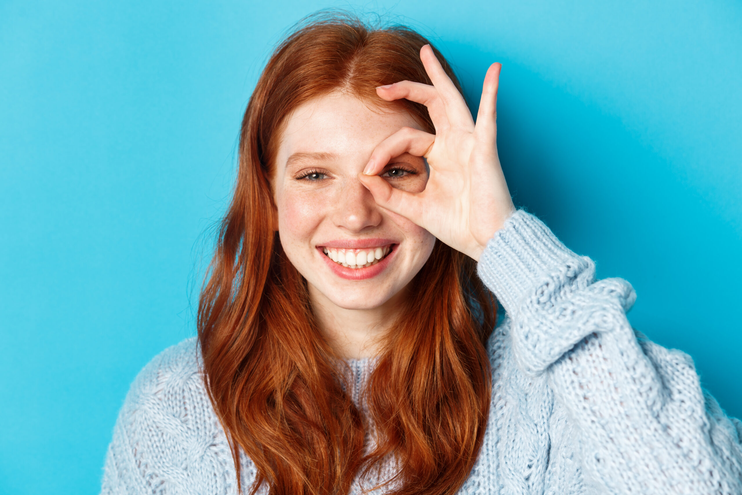 Headshot of cheerful redhead female model showing okay sign over eye, smiling satisfied and happy, standing against blue background Headshot of cheerful redhead female model showing okay sign over eye, smiling satisfied and happy, standing against blue background