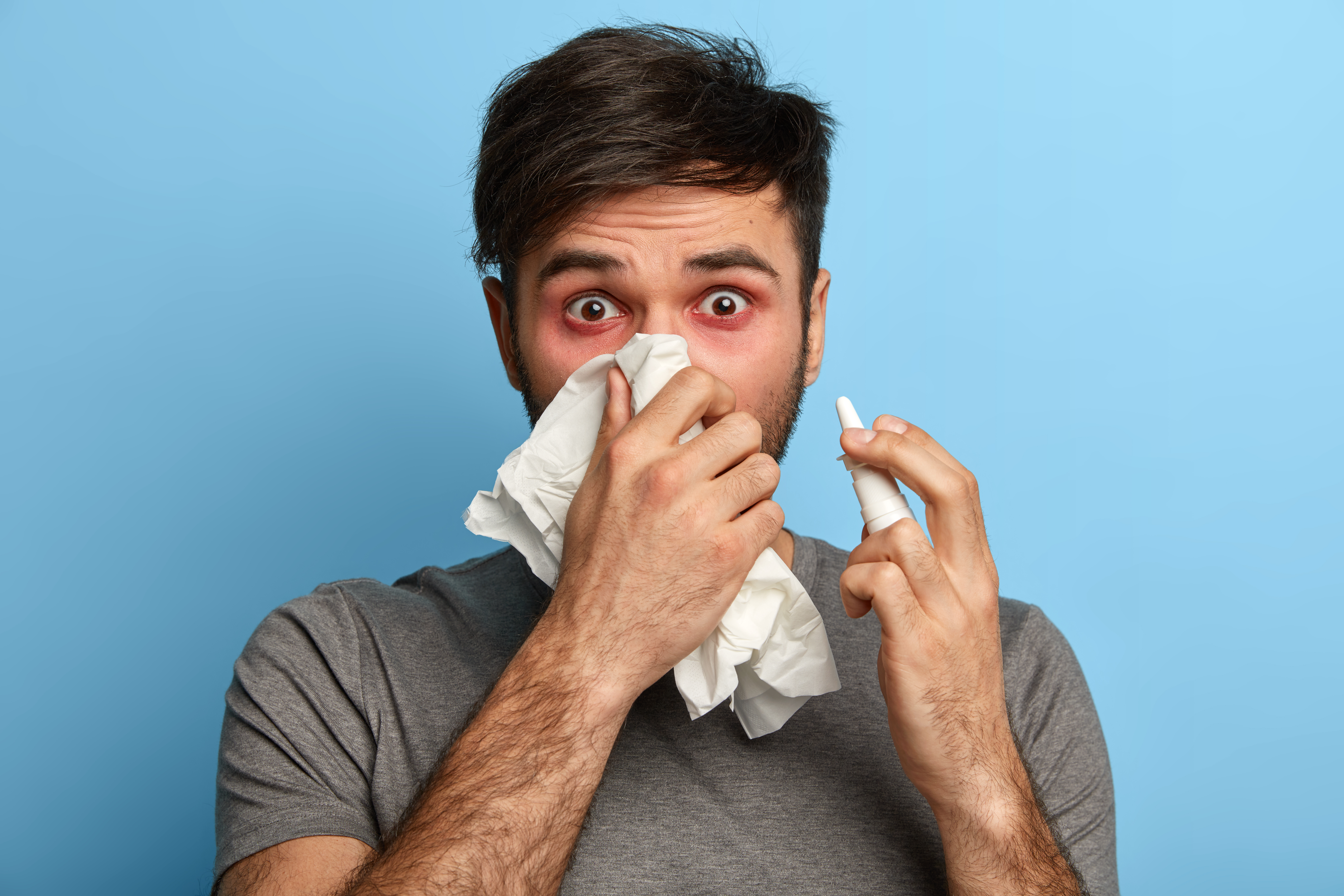 Health care., flu, allergy and hygiene concept. Sick tired man blows nose in paper napkin, uses nasal spray as has rhinitis, sneezes constantly, has symptoms of influenza, fever or coronavirus Health care., flu, allergy and hygiene concept. Sick tired man blows nose in paper napkin, uses nasal spray as has rhinitis, sneezes constantly, has symptoms of influenza, fever or coronavirus