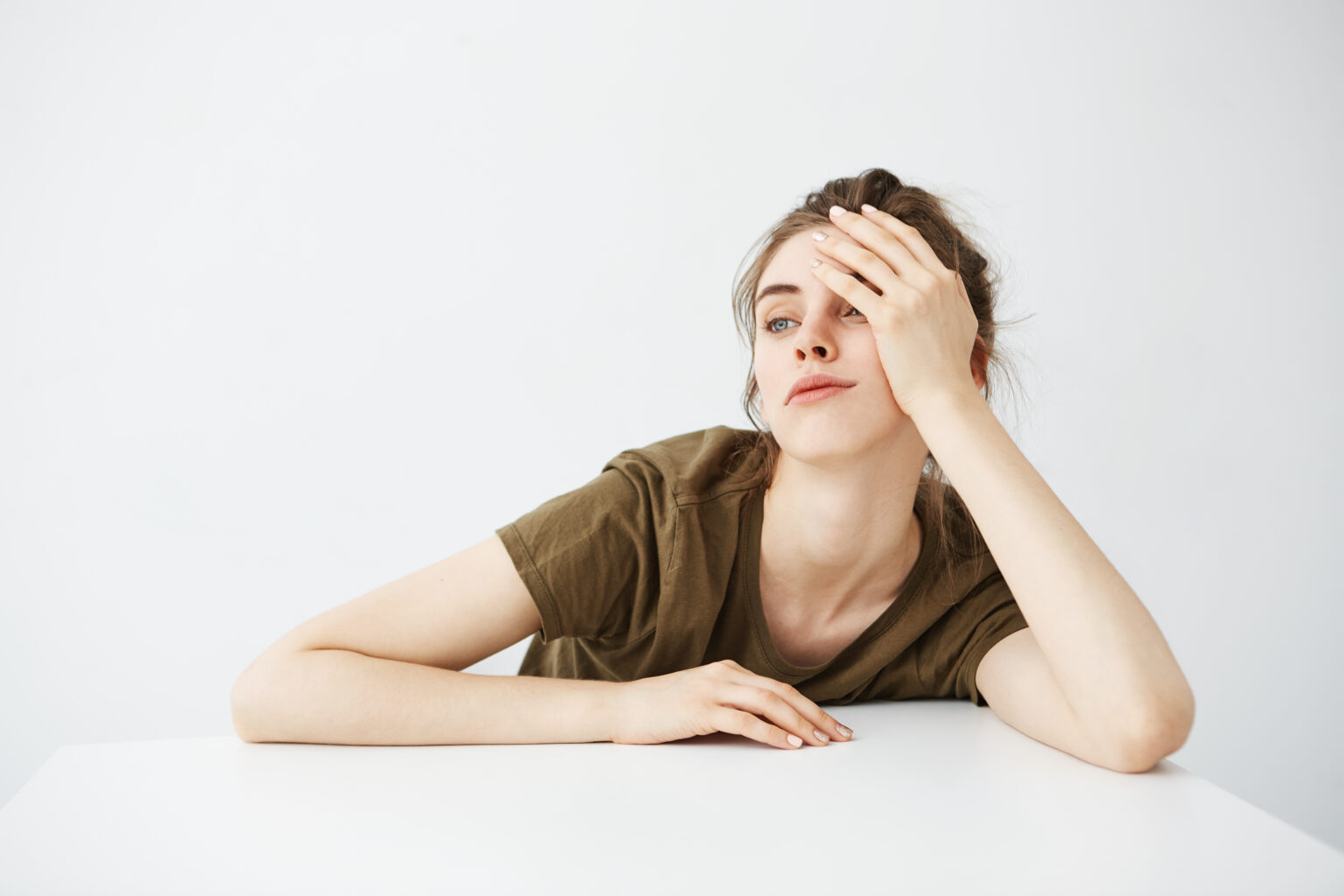 Bored tired dull young girl student with bun sitting at table over white background. Bored tired dull young girl student with bun sitting at table over white background.