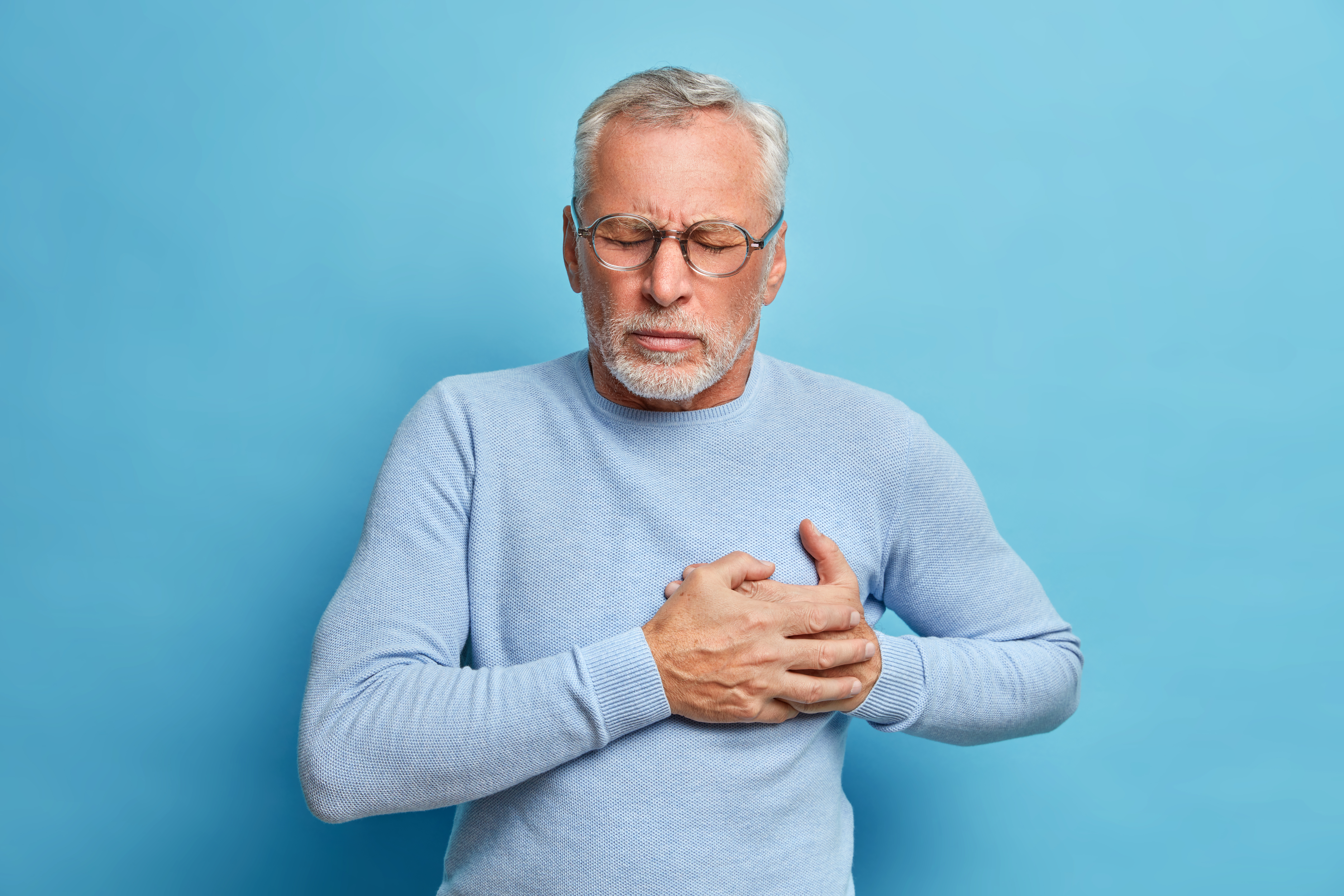 Senior man in spectacles presses hand to chest has heart attack suffers from unbearable pain closes eyes wears optical glasses poses against blue background. People age and problems with health Senior man in spectacles presses hand to chest has heart attack suffers from unbearable pain closes eyes wears optical glasses poses against blue background. People age and problems with health