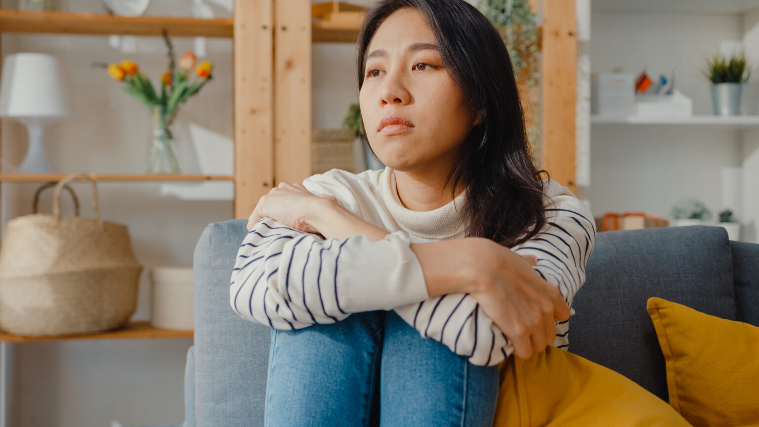 Thoughtful Asia lady sit embracing knees at sofa in living room