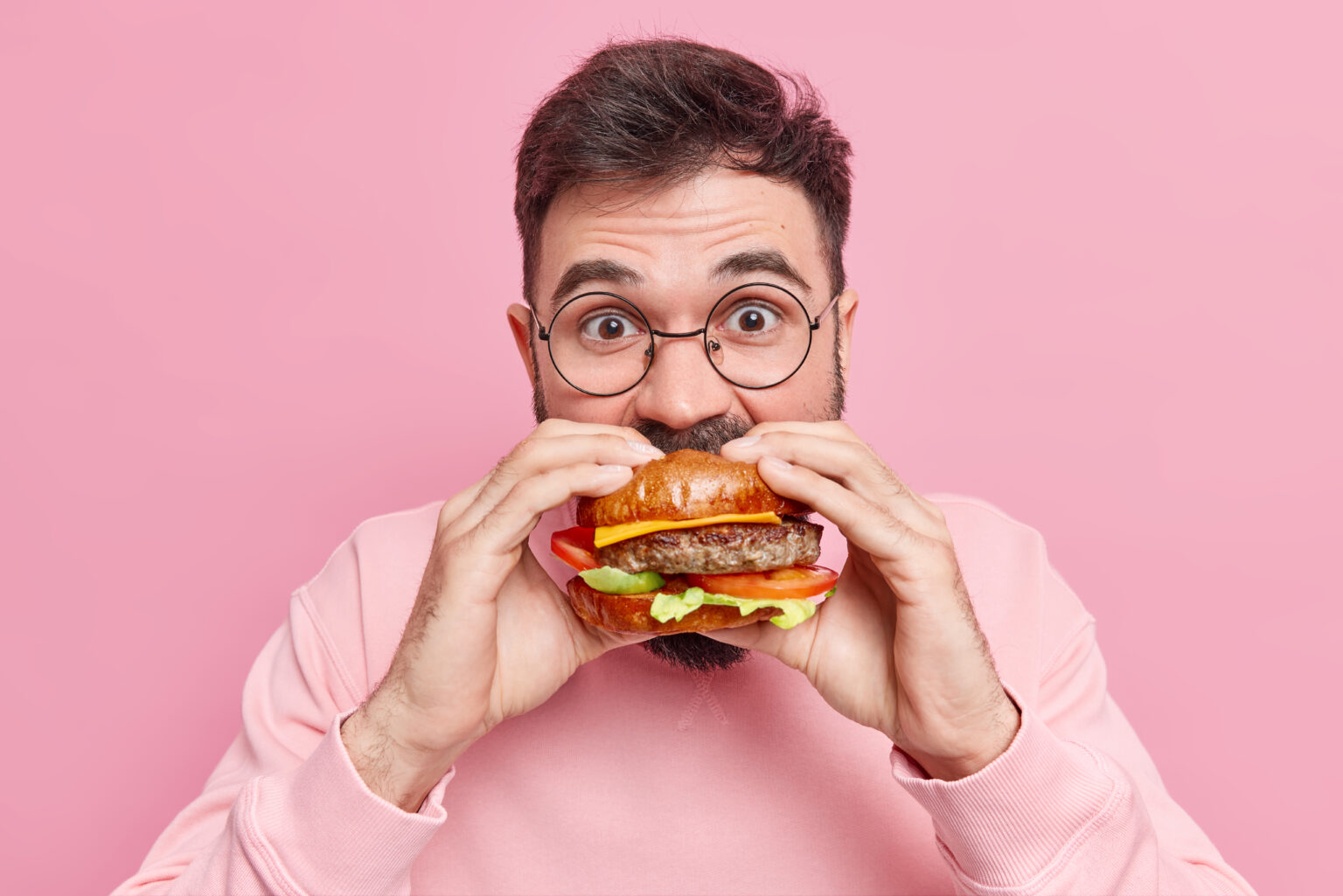 Close up shot of young man eats greedily delicious hamburger feels very hungry consumes fast food wears round spectacles and jumper isolated over pink background. Unhealthy nutrition concept Close up shot of young man eats greedily delicious hamburger feels very hungry consumes fast food wears round spectacles and jumper isolated over pink background. Unhealthy nutrition concept