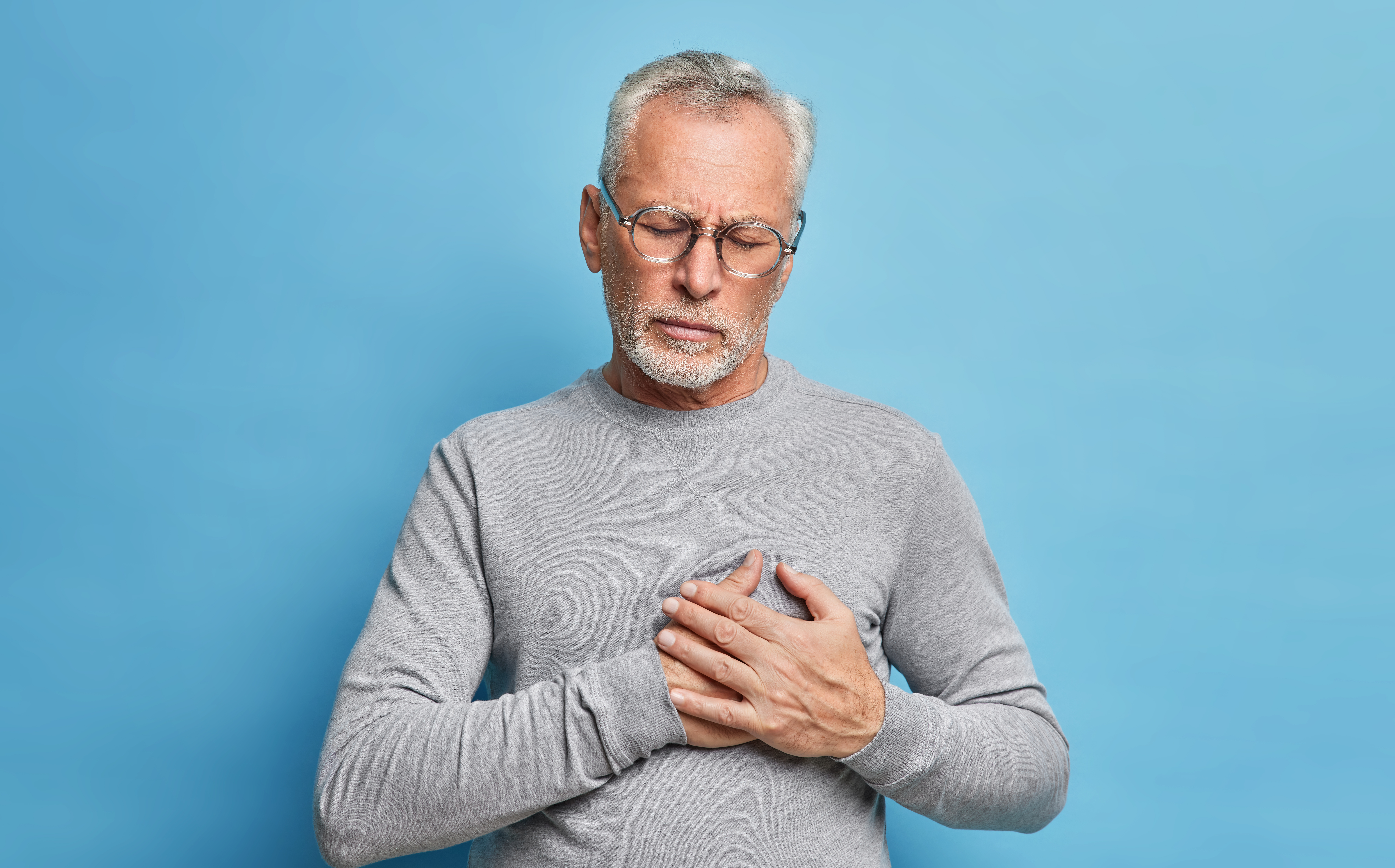 Indoor shot of senior bearded man has heart attack suffers from painful feelings needs painkillers presses hands to chest being unhealthy wears spectacles and grey casual jumper isolated on blue wall