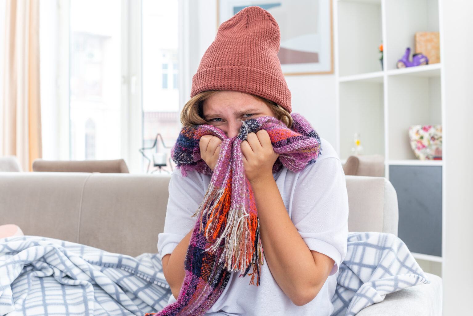 unhealthy young woman in warm hat with scarf around neck feeling unwell and sick suffering from cold and flu looking worried sitting on couch in light living room unhealthy young woman in warm hat with scarf around neck feeling unwell and sick suffering from cold and flu looking worried sitting on couch in light living room