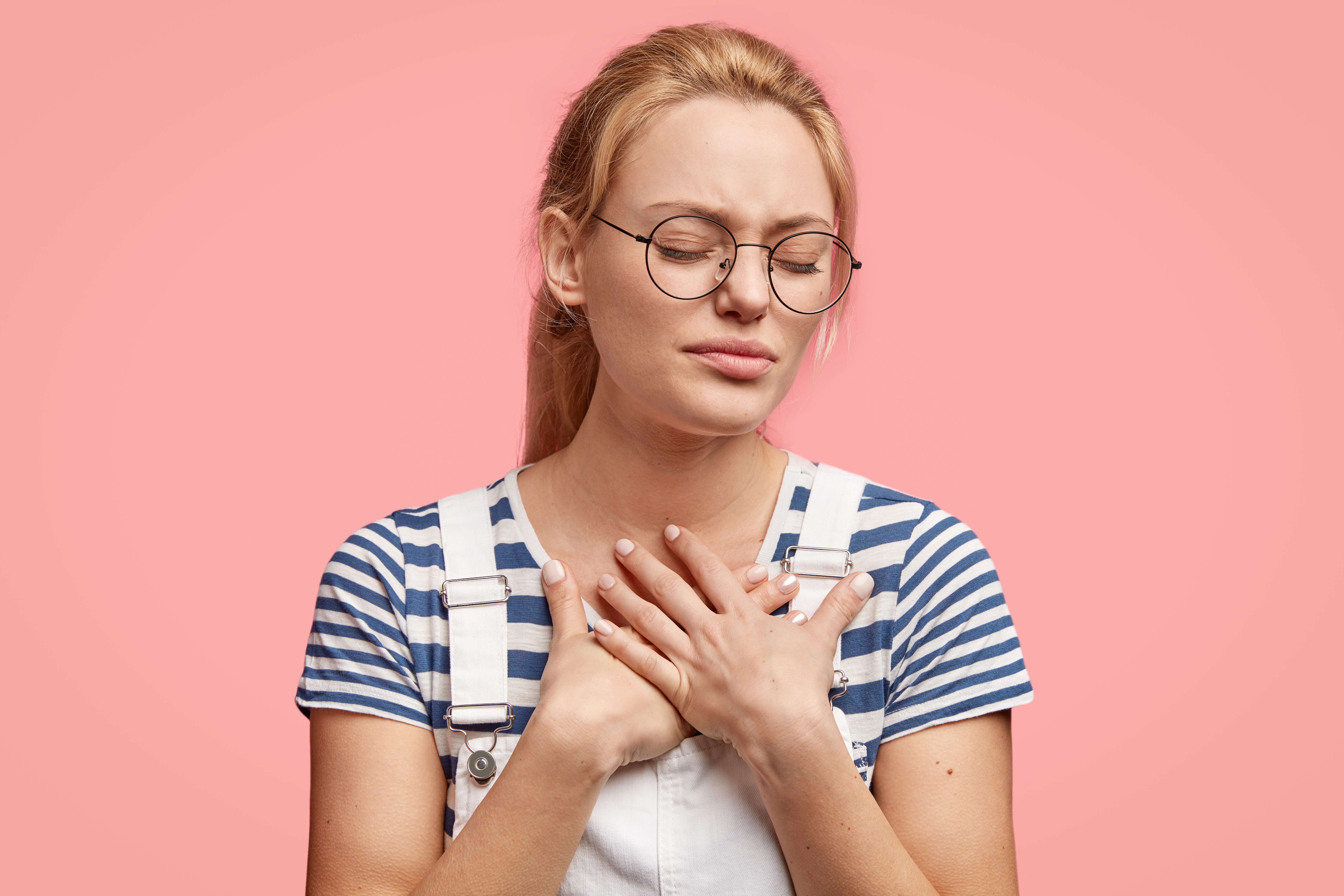 Sad stressful young Caucasian female keeps hands on chest, feel pain in heart, closes eyes, stands against pink background. Beautiful woman recieves bad news. Negative facial expressions concept Sad stressful young Caucasian female keeps hands on chest, feel pain in heart, closes eyes, stands against pink background. Beautiful woman recieves bad news. Negative facial expressions concept