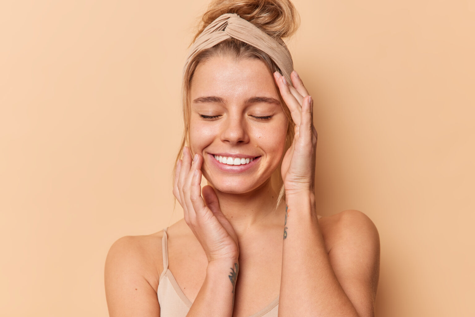 Photo of pleased young European woman keeps eyes closed touches face gently has natural beauty healthy clean skin wears headband has combed hair isolated over beige background. Wellness concept Photo of pleased young European woman keeps eyes closed touches face gently has natural beauty healthy clean skin wears headband has combed hair isolated over beige background. Wellness concept