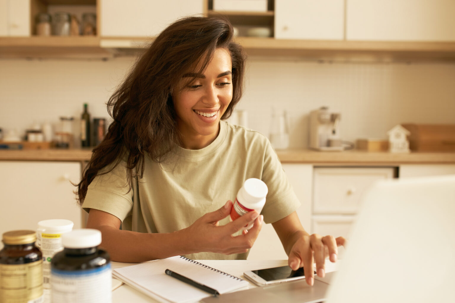 Biohacking and superfoods concept. Cheerful young Latin female nutritionist holding bottle of dietary supplements for muscle building. Pretty girl using laptop ordering vitamins via online store Biohacking and superfoods concept. Cheerful young Latin female nutritionist holding bottle of dietary supplements for muscle building. Pretty girl using laptop ordering vitamins via online store