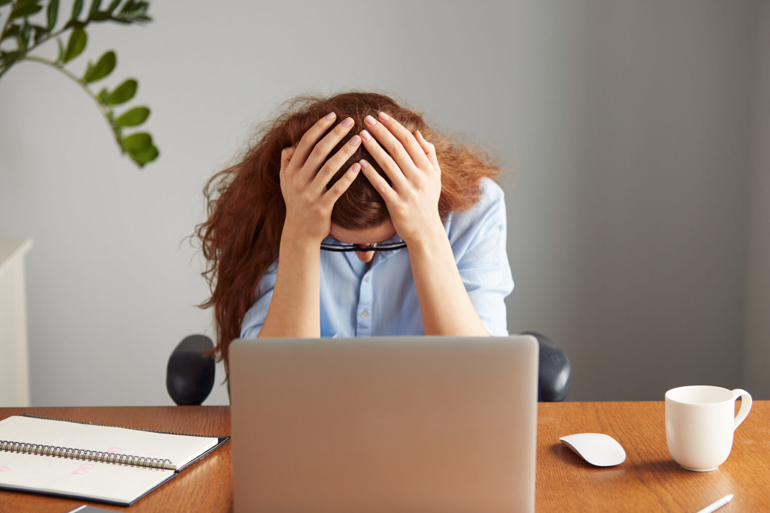 Headshot of exhausted redhead female office worker in blue shirt and glasses sitting in front of the laptop at the wooden office desk, holding her head in despair. Overwork and deadline concept Headshot of exhausted redhead female office worker in blue shirt and glasses sitting in front of the laptop at the wooden office desk, holding her head in despair. Overwork and deadline concept
