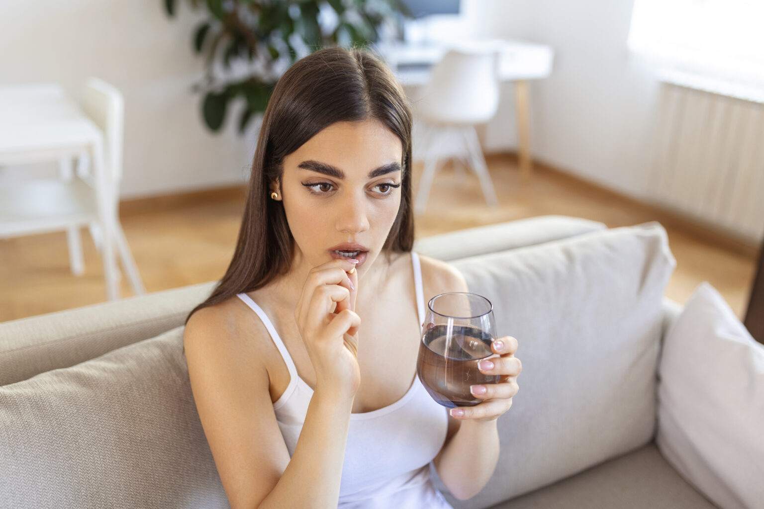 Concept of suffering from terrible pain. Beautiful, sad, upset, unhappy, troubled, weak woman with black hair, wearing casual clothes is sitting on a sofa and taking a pill Concept of suffering from terrible pain. Beautiful, sad, upset, unhappy, troubled, weak woman with black hair, wearing casual clothes is sitting on a sofa and taking a pill