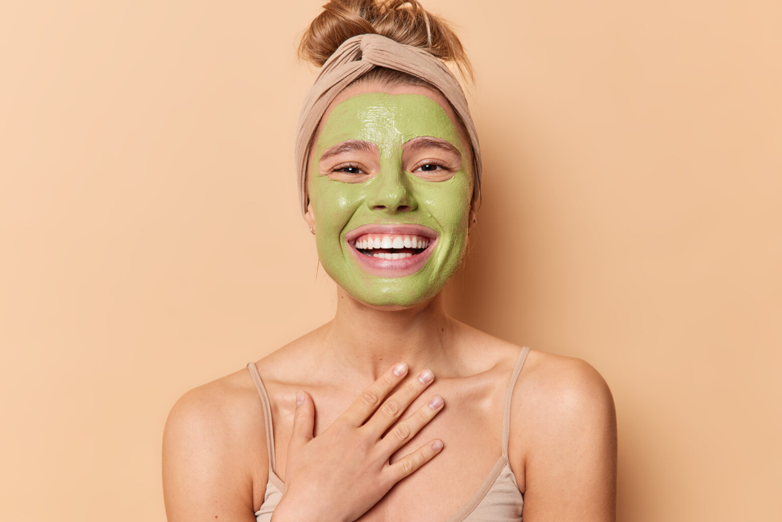 Happy European woman smiles broadly keeps hand on chest applies nourishing green mask undergoes beauty treatmentss wears headband has combed hair isolated over brown background. Skin care concept Happy European woman smiles broadly keeps hand on chest applies nourishing green mask undergoes beauty treatmentss wears headband has combed hair isolated over brown background. Skin care concept
