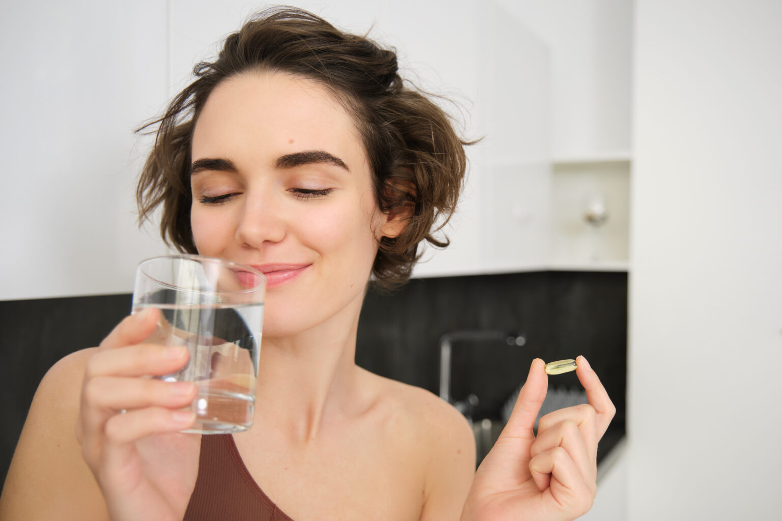Portrait of sportswoman drinking water, taking vitamins, dietry supplements for healthy skin, having omega-3 pill, standing in her kitchen in workout clothing Portrait of sportswoman drinking water, taking vitamins, dietry supplements for healthy skin, having omega-3 pill, standing in her kitchen in workout clothing
